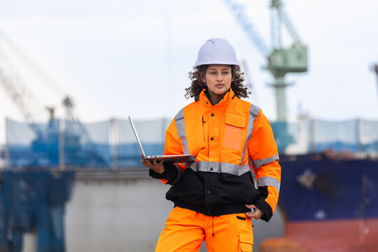 Professional female engineer holding laptop monitoring shipping operations with digital equipment at harbor, Portrait of a female site supervisor in high visibility uniform working at commercial port