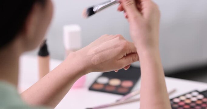 Woman applying foundation onto her hand with makeup brush at table indoors, closeup