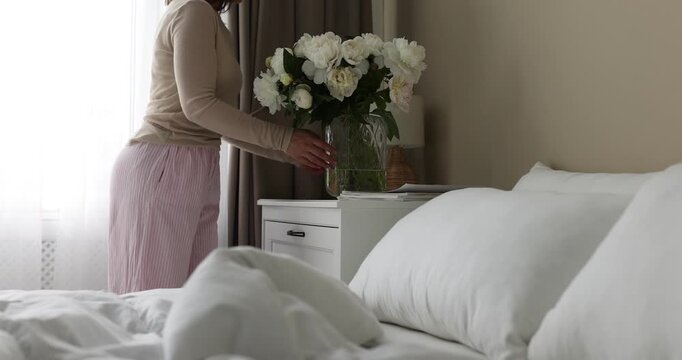 Woman putting vase with beautiful peonies on bedside table and turning on lamp at home, closeup