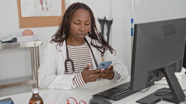 Woman doctor with stethoscope scrolling smartphone at clinic desk with computer, prescription bottle, paperwork and crutches visible; concentration care professionalism.