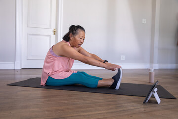 Senior Asian woman stretching forward on exercise mat at home, wearing pink top, following tablet © wavebreak3