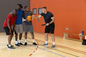 Naklejka premium Diverse male basketball unit standing on gym court holding orange basketball, coach pointing tablet