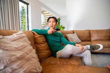 Obraz premium Asian man sitting on brown leather sectional wearing green sweater holding black TV remote at home