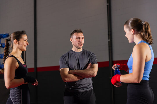 Male trainer in sportswear instructing two female trainees in boxing ring corner, wraps and bottle