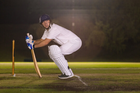 Wooden cricket bat standing vertically in marked crease with stumps on grass under floodlights