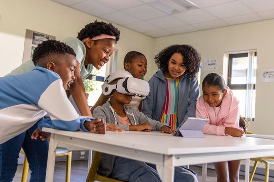 Diverse preteen children gathering at table at school watching peer with VR headset using tablet