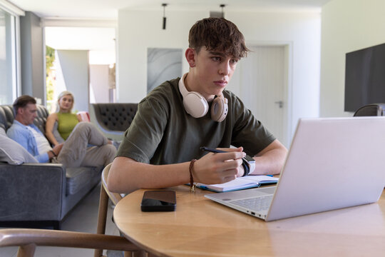 Family with teen son sitting at table using laptop headphones while parents using smartphone