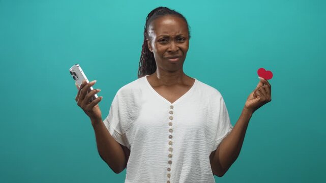 Young woman holding smartphone in left hand and pinching red paper heart with right fingers in studio; skepticism.