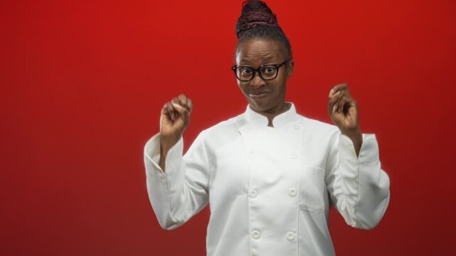 Woman chef wearing white uniform and glasses makes air quotes with hands against a red studio backdrop, bun hair visible; playful skepticism.