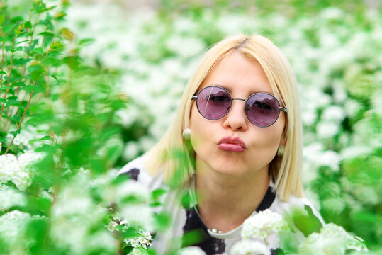 blonde girl in round glasses makes a kiss gesture with her lips against a background of white flowers