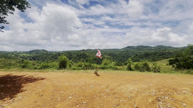 A Puerto Rican flag stands on a simple pole along a red dirt road, with rolling green hills and dense tropical vegetation stretching across the background. Large white clouds fill the blue sky above, 