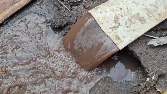 A high-angle view of brown, polluted wastewater flowing rapidly and bubbling out of a large canvas hose onto the sandy ground.