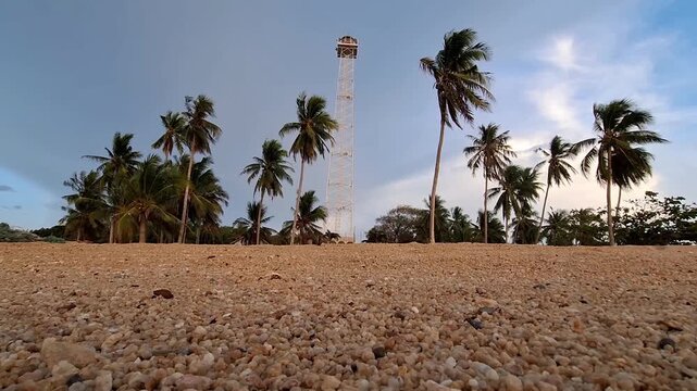 A low-angle shot of a tall white steel skeleton lighthouse tower surrounded by tropical coconut palm trees on a coarse pebble sand beach.