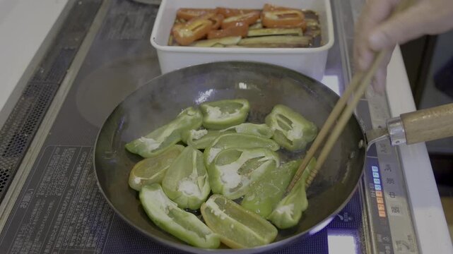 煮浸しで緑色のピーマンを鉄フライパンの中で裏返しし、IHクッキングヒーターの中火で焼き上げるシーン