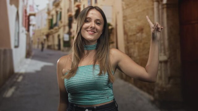 Woman raises bare arm and points finger upward on a narrow street wearing turquoise crop top and choker; effortless confidence.