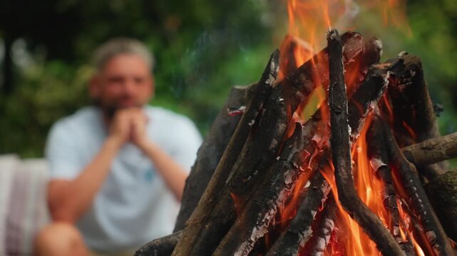 Close-up of burning campfire with orange flames and stacked wooden logs in sharp focus. Blurred man in white shirt sits contemplatively in background with green forest bokeh.