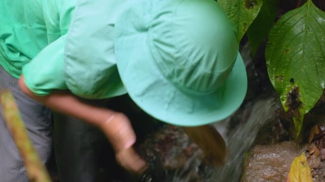 Person in mint green outdoor clothing and matching bucket hat crouches beside a small jungle stream, washing hands in cool water as it cascades over mossy rocks amid tropical foliage and fallen.