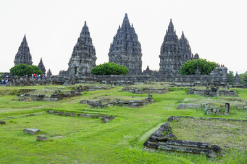 Obraz premium Ancient temple towers rise above green grass. Stone fragments lie scattered across the earth. Visitors explore the ruins with quiet reverence. History whispers through weathered stone and lush grass