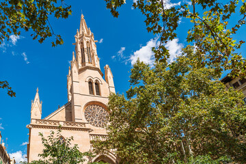 Fototapeta premium Bellver Church in Palma de Mallorca, Mallorca, displays Gothic architecture with its tower and rose window beneath a clear blue sky, framed by green trees and appealing to sun.