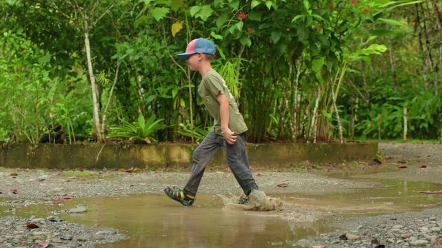 Boy happily jumps through muddy puddle in slow motion. Child wearing colorful cap splashes water on gravel path surrounded by lush green tropical plants and foliage after rain.