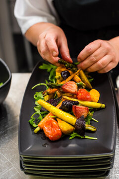 Chef's hands preparing roasted vegetable salad including tomato, carrot and asparagus on plate, close up