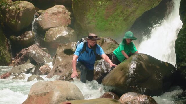 Father and son adventure through lush tropical rainforest, carefully climbing over wet mossy boulders beside a rushing waterfall, mist filling the air as they explore the scenic river canyon together.