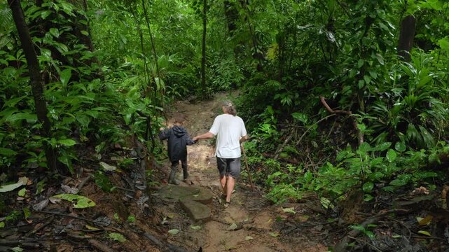 Adult man and young boy walk together on muddy path through lush green tropical rainforest. Father holds walking stick while guiding child through dense jungle vegetation.