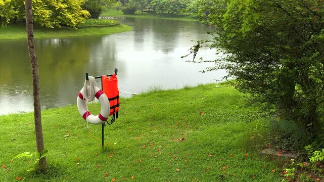 A tranquil, static shot of a serene public park during a gentle rain. In the foreground, a red and white lifebuoy ring, an orange life jacket