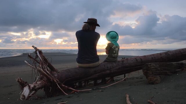 Father and young child wearing hats sit on weathered driftwood log on dark sandy beach, gazing at golden sunset breaking through dramatic cloudy sky over calm ocean waves at twilight.
