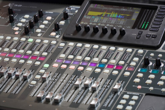 Close-up of a professional digital audio mixing console with illuminated colorful LED buttons, knobs, and channel faders in a dark music recording studio or broadcasting control room.