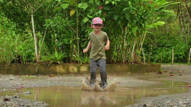 Boy happily jumps through muddy puddle in slow motion creating dramatic water splashes. Child wearing cap plays outdoors in tropical setting with lush green foliage in background after rain.