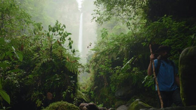 Son with father hiking through wild nature in lush tropical rainforest. Man with walking stick and backpack navigates mossy rocks as tall waterfall cascades through misty green jungle foliage.