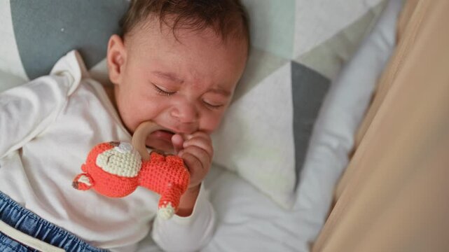 Hispanic boy chewing a crocheted orange fox teether on a wooden ring, gripping it with hand in crib inside building; teething discomfort soothing.