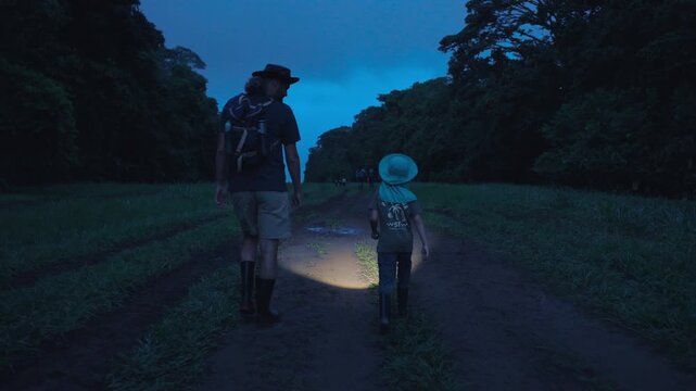 Father and child wearing hats and rubber boots trek along a muddy dirt path at dusk, using flashlights to illuminate their way through lush green tropical forest under a moody blue twilight sky.