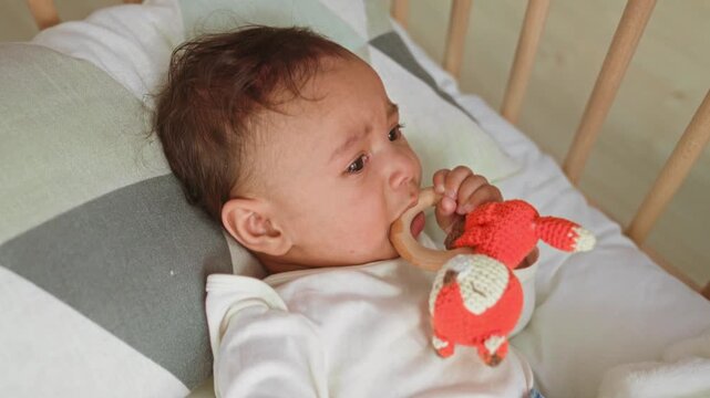 Baby boy in white onesie bites wooden teether ring with his hand and cries on a pillow in a nursery crib; teething discomfort.