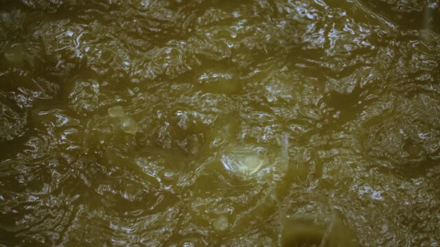 Close-up view of murky olive-green water surface with rippling texture and small bubbles rising through turbulent flow, creating abstract organic pattern with light reflections dancing across liquid.