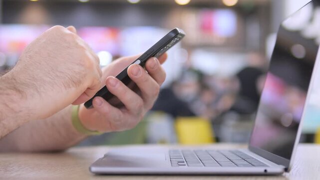 Hand holds smartphone near open laptop. Focus on fingers scrolling device. Background shows public space blur. Person seems concentrated, multitasking. Ideal for business, work, or tech themes