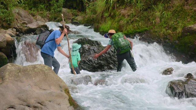 Three hikers with backpacks carefully wade through fast-flowing mountain stream using wooden walking sticks for balance. Lush green vegetation and mossy rocks surround the white water rapids.