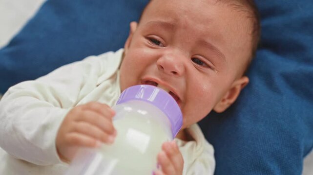 Hispanic baby boy crying, grasping milk bottle with tiny hand to open mouth on bed pillow in bedroom, tears visible and face scrunched; distress hungry.