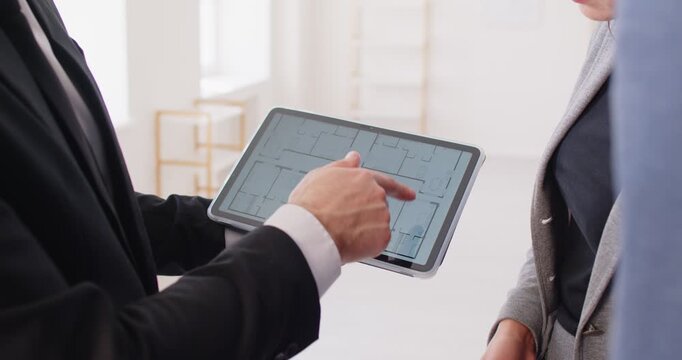 Unrecognizable male broker meeting together with client, looking at project plan, showing blueprints on digital tablet, planning work. Two architects with blueprint construction project