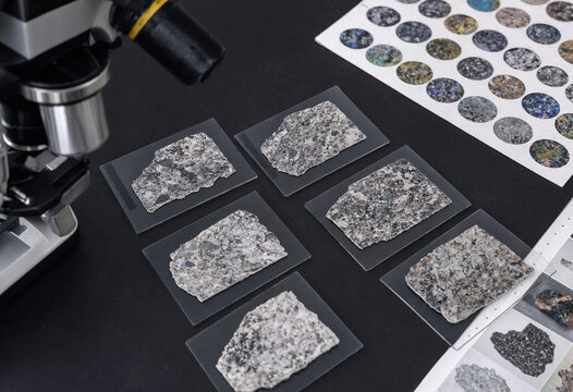 Gray stone rock fragments on glass slides next to a microscope and a circular color chart on a dark laboratory table for geological study