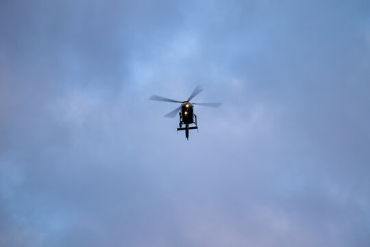 Police helicopter in flight viewed from below against an overcast blue-grey sky with searchlight illuminated