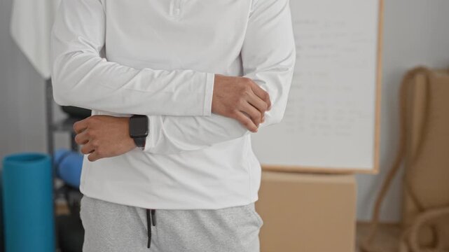 Man holding forearm with smartwatch, wearing white long sleeve and gray sweatpants, stretching beside yoga mat, foam roller and exercise ball in studio; concentration.