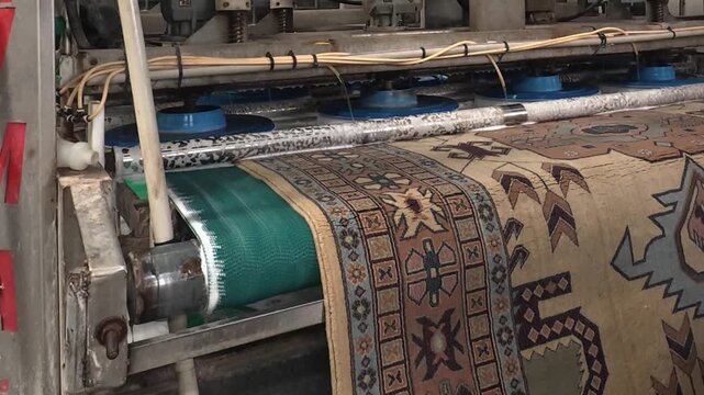 A patterned carpet moves through industrial rollers while a worker applies liquid in a bright factory setting, showing textile processing, maintenance