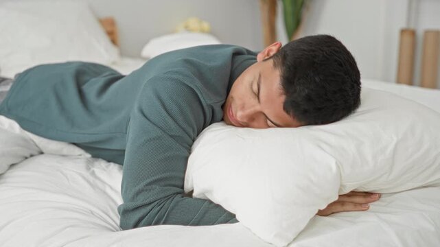 Man lying prone on bed, sleeping face on a white pillow with arm tucked under pillow in a studio bedroom set wearing a green sweater; calm rest.
