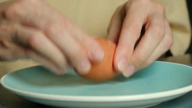 Human hands cracking, carefully peeling shell off hard boiled brown egg over blue plate, simple process for preparing healthy protein rich meal or snack in kitchen. Hands peeling boiled egg over plate