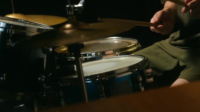 Professional drummer playing powerful beat on drum kit in dark studio setting. Musician strikes snare drum and cymbals with wooden drumsticks during live performance. Drummer playing drum kit