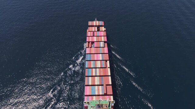 Stunning top-down aerial shot following a massive cargo ship loaded with colorful intermodal containers as it powerfully sails through the deep blue ocean, representing global trade and logistics