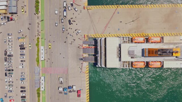 Top-down aerial view showing cars, trucks, and passengers boarding a large ro-ro ferry docked at the pier, highlighting the bustling activity of maritime transport and logistics in a harbor