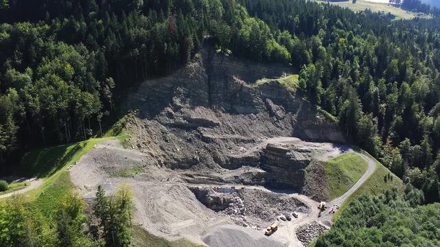 Aerial view of an active quarry surrounded by forest. Exposed rock faces, excavation terraces, gravel stockpiles, access roads, and heavy machinery highlight industrial mineral extraction operations.